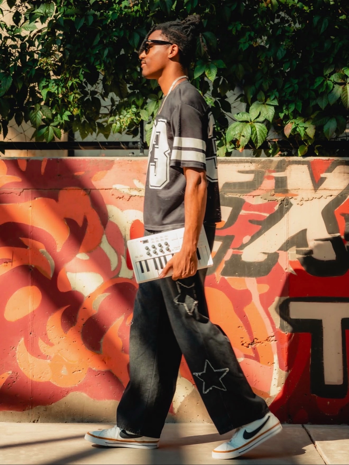 Jaywill Williams walking past a graffiti wall holding a MIDI keyboard, wearing a number 33 jersey and sunglasses